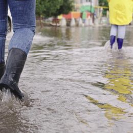 two people walk through puddles of flooded city rubber boots on their feet, teamwork, girls summer raincoats, lot of rainwater is poured on street, natural weather phenomena, walking boots on road
