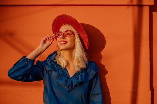 Happy smiling fashionable woman wearing trendy color sunglasses, hat, blue dress, posing outdoor, near orange wall during the sunset. Copy, empty space for text
