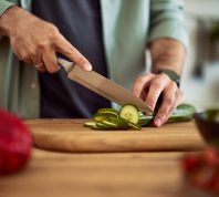A close-up shot of male hands using a sharp kitchen knife to cut a fresh cucumber for a salad on a wooden cutting board.