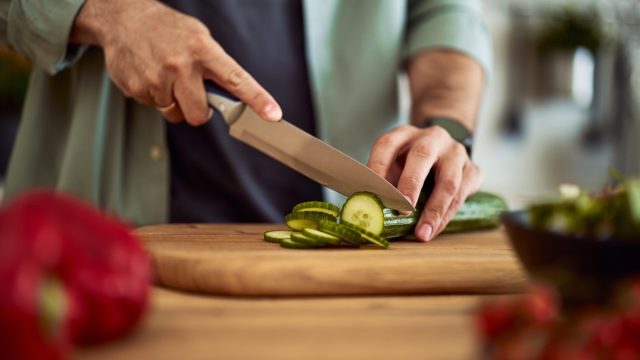 A close-up shot of male hands using a sharp kitchen knife to cut a fresh cucumber for a salad on a wooden cutting board.