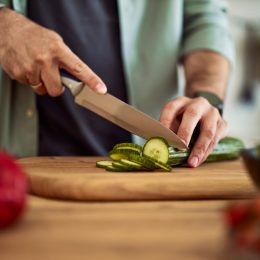 A close-up shot of male hands using a sharp kitchen knife to cut a fresh cucumber for a salad on a wooden cutting board.