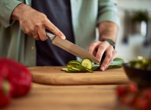 A close-up shot of male hands using a sharp kitchen knife to cut a fresh cucumber for a salad on a wooden cutting board.