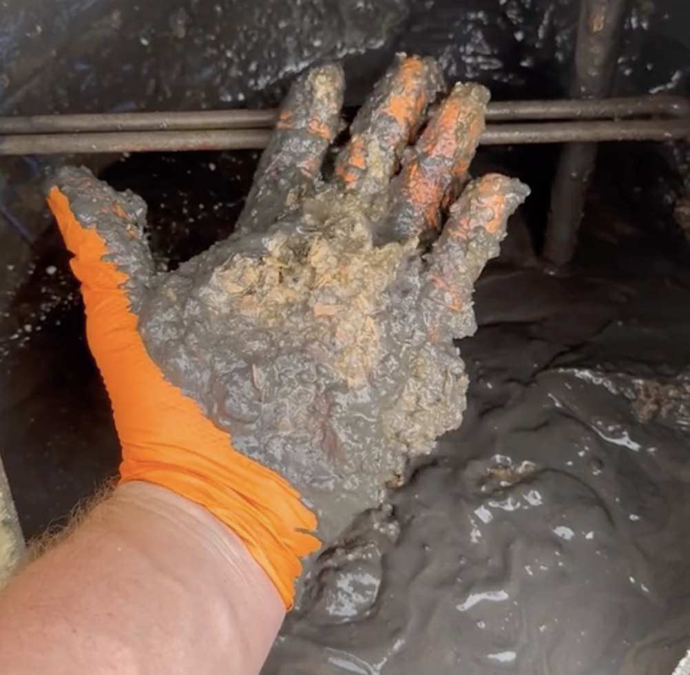 Still from a video showing a hand wearing an orange glove, pulling black gunk out of a hot water heater
