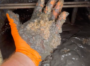 Still from a video showing a hand wearing an orange glove, pulling black gunk out of a hot water heater