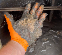 Still from a video showing a hand wearing an orange glove, pulling black gunk out of a hot water heater