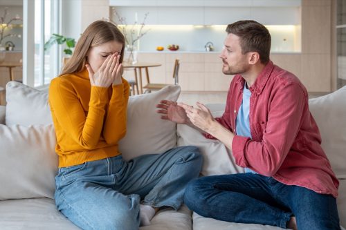 Jealous furious man talk scream at crying woman wife sitting on couch at home. Family quarrel, scandal of two spouses. Tyrant husband threatens woman. Betrayal infidelity mistrust, couple relations.