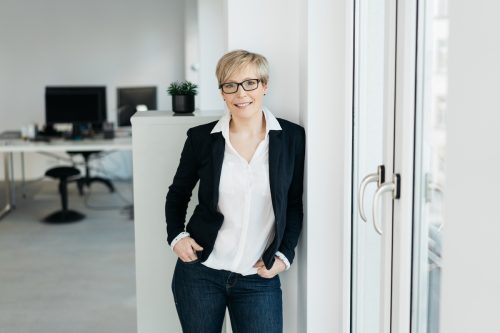 Woman wearing a white blouse, black blazer, and jeans leaning against a wall in an office