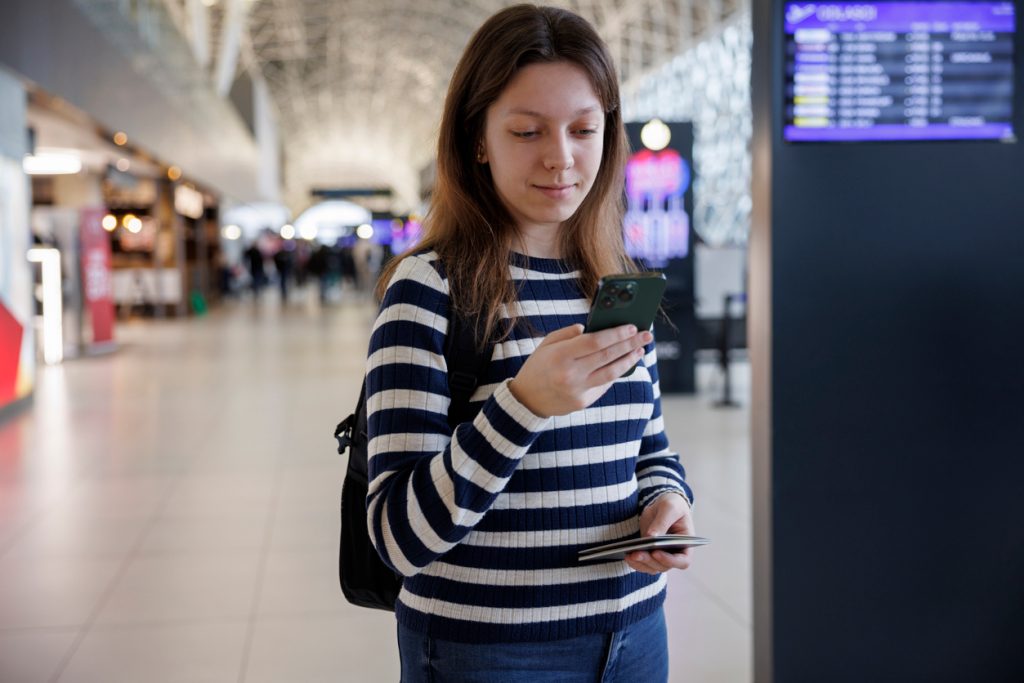 A woman standing in the airport holding her phone and passport