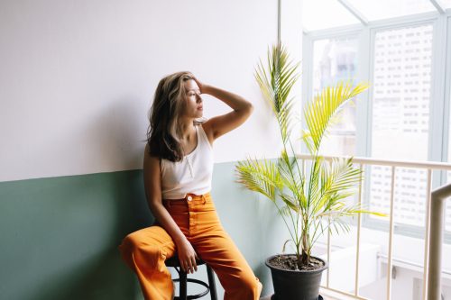 A woman wearing orange pants and white tank top sits on a stool next to a houseplant while looking out a window