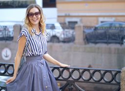 young woman walks in Saint-Petersburg; she's smiling and posing near a railing wearing a blue and white striped blouse and a blue skirt