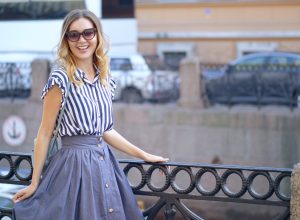young woman walks in Saint-Petersburg; she's smiling and posing near a railing wearing a blue and white striped blouse and a blue skirt