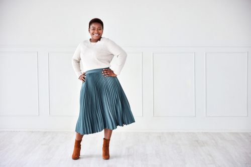 confident woman posing in front of a white wall wearing a pleated, teal A-line skirt, white sweater, and brown booties