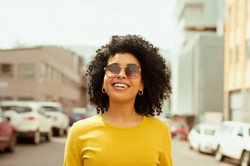Shot of a young woman looking happy while out in the city; she's wearing a yellow shirt and sunglasses
