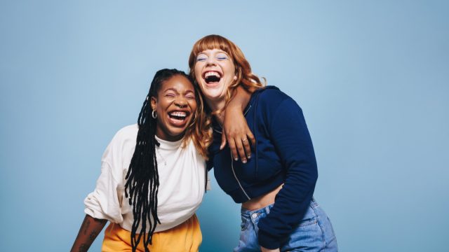 two young women laughing with arms around each other on a blue background