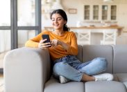Happy Young Female Messaging On Smartphone While Relaxing On Couch At Home, Smiling Eastern Woman Using Mobile Phone, Texting With Friends Or Browsing New App For Online Shopping, Copy Space