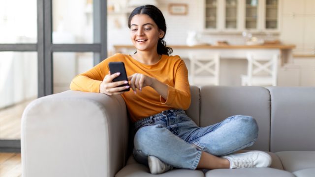 Happy Young Female Messaging On Smartphone While Relaxing On Couch At Home, Smiling Eastern Woman Using Mobile Phone, Texting With Friends Or Browsing New App For Online Shopping, Copy Space