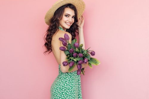 Side view of a young woman wearing a backless green dress and a straw hat, holding purple tulips, against a pink background