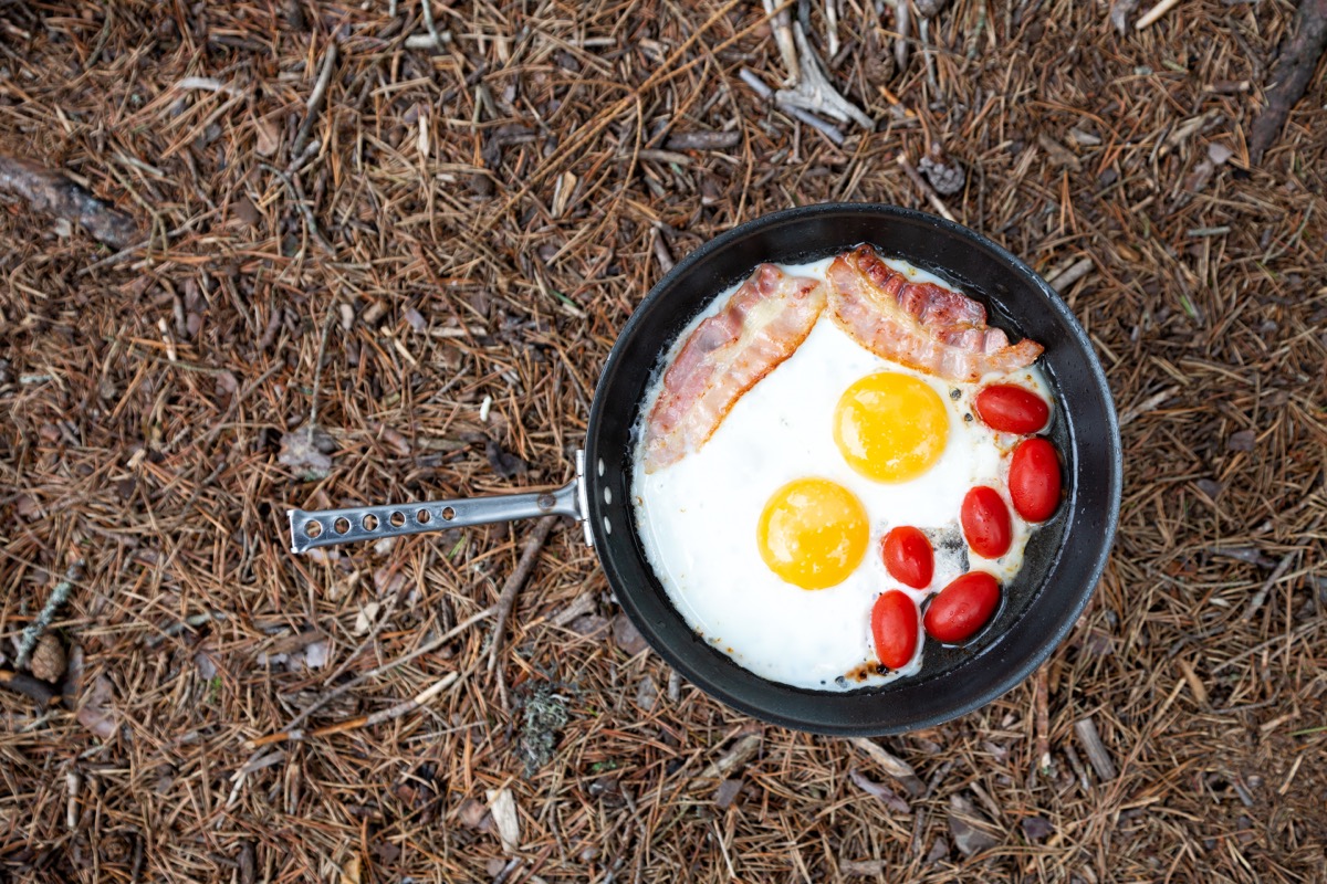 Frying pan with fried eggs, bacon and tomatoes in nature. Cooking breakfast on a gas stove at a summer camp.