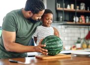 Man and little boy standing at kitchen counter with whole watermelon on cutting board