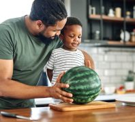 Man and little boy standing at kitchen counter with whole watermelon on cutting board