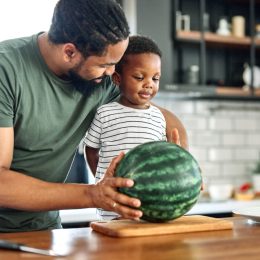 Man and little boy standing at kitchen counter with whole watermelon on cutting board