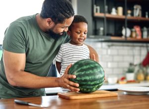 Man and little boy standing at kitchen counter with whole watermelon on cutting board