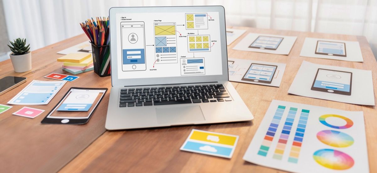 Digital and paper wireframe designs for web or mobile app UI UX display on laptop computer screen. Panoramic shot of developer workspace for brainstorming and design application framework. Scrutinize