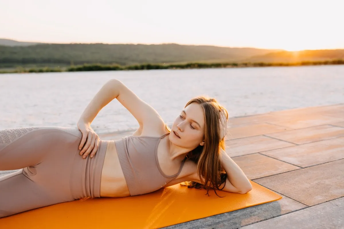 Young slim fit woman on a sport mat in yoga side lying pose with leg lift, at the sunset, outdoors. Pilates teacher doing exercises.