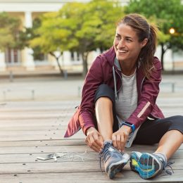 Mature fitness woman tie shoelaces on road. Cheerful runner sitting on floor on city streets with mobile and earphones wearing sport shoes. Active latin woman tying shoe lace before running.