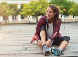 Mature fitness woman tie shoelaces on road. Cheerful runner sitting on floor on city streets with mobile and earphones wearing sport shoes. Active latin woman tying shoe lace before running.