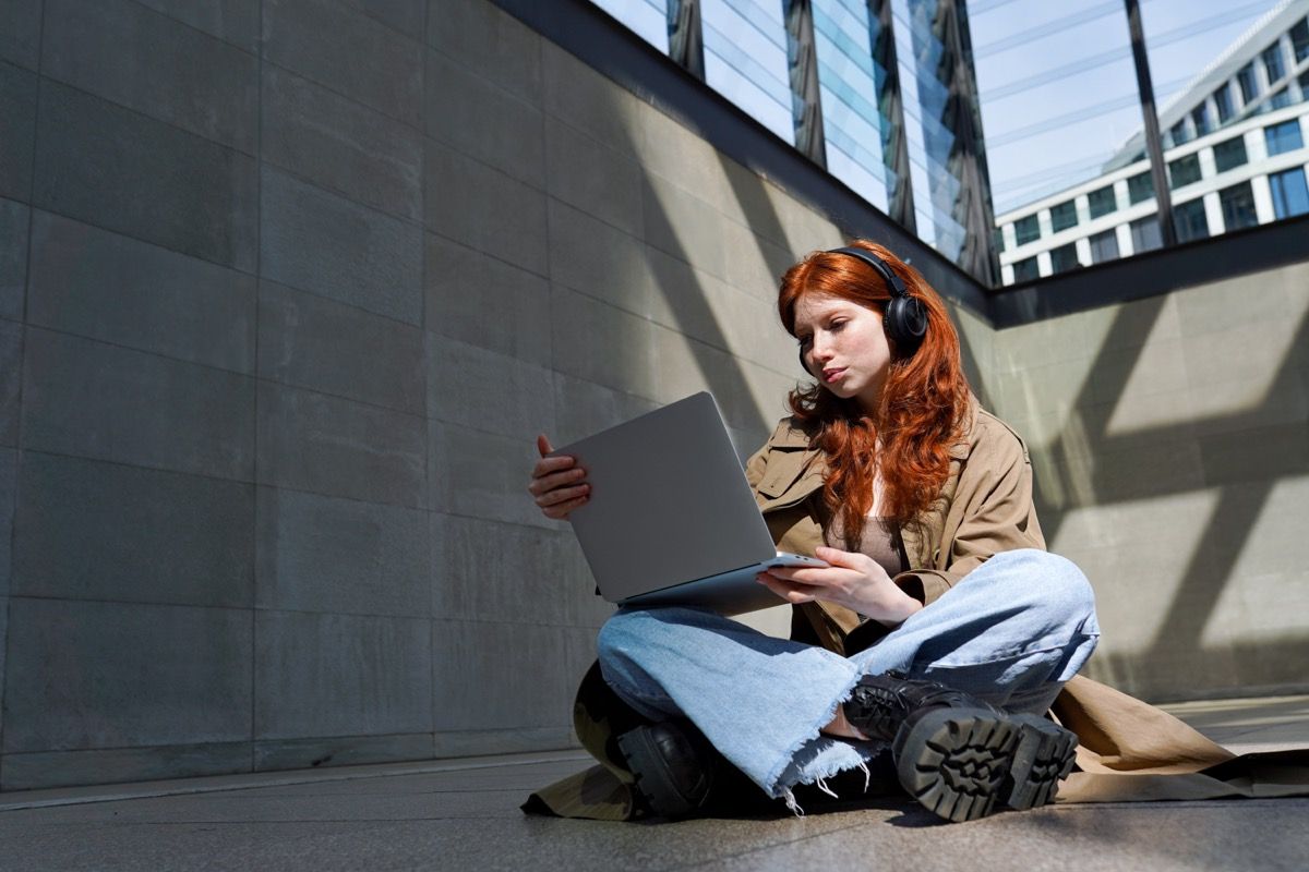 Redhead hipster girl student wearing headphones using laptop in city urban location searching information, online learning, watching webinar class or having video chat outdoors.