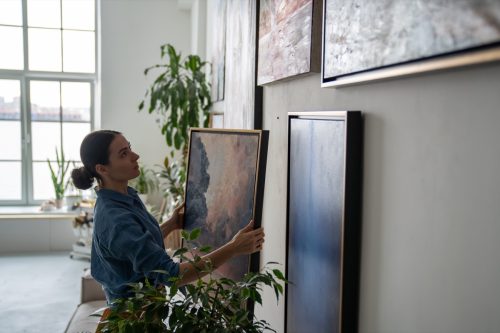 Thoughtful female artist holding painting in frame, hanging on wall. Serious girl creating colorful composition of paintings at apartments, making cozy creative atmosphere in home art studio workshop.