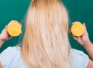 back of a blonde woman holding two halfs of a lemon on a dark green background