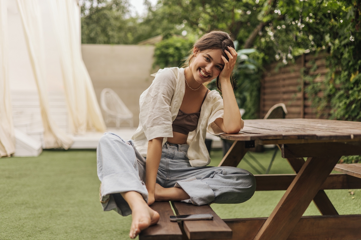 young woman with beaming smile looks at camera while sitting at a picnic table. She is wearing summery linen clothes