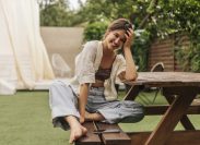 young woman with beaming smile looks at camera while sitting at a picnic table. She is wearing summery linen clothes