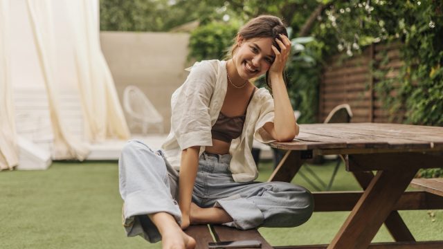 young woman with beaming smile looks at camera while sitting at a picnic table. She is wearing summery linen clothes