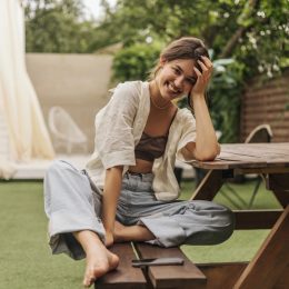 young woman with beaming smile looks at camera while sitting at a picnic table. She is wearing summery linen clothes