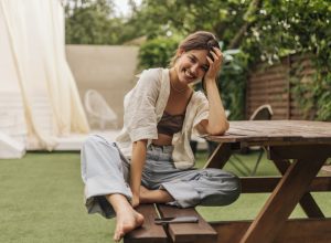 young woman with beaming smile looks at camera while sitting at a picnic table. She is wearing summery linen clothes