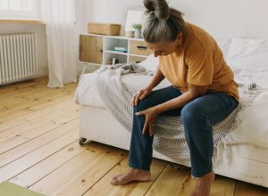 Elderly woman have pain in knee. Mature female suffer with trick knee sitting on bed at home, cozy bedroom interior. Growing old, take care of health, health problems concept