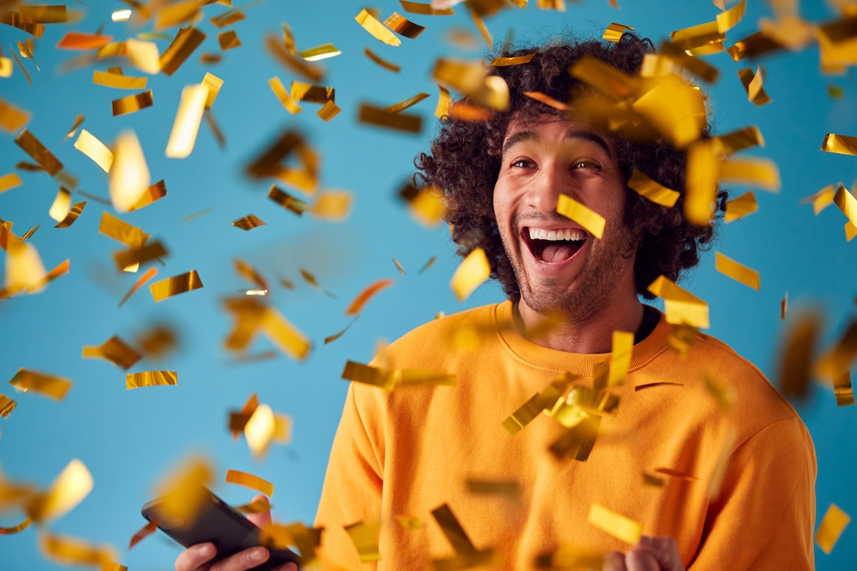Young man in a yellow sweatshirt celebrating against a teal blue background with gold confetti everywhere