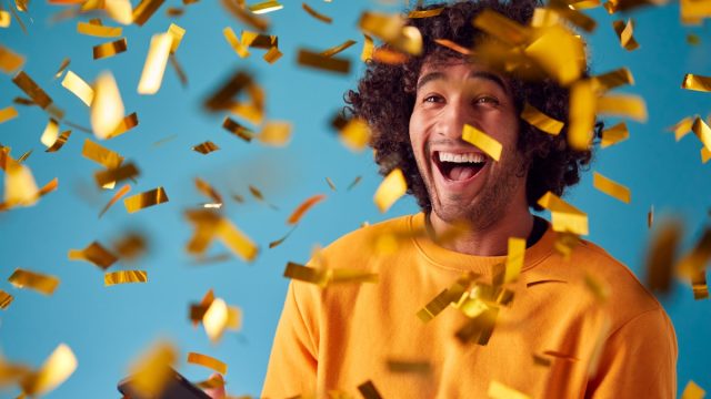Young man in a yellow sweatshirt celebrating against a teal blue background with gold confetti everywhere
