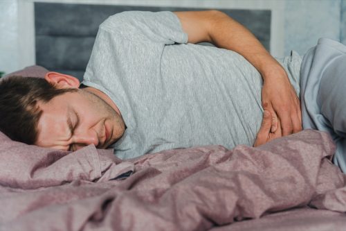 Frustrated handsome young man hugging his belly suffering from stomach pain while lying in bad at home. Male with belly ache. Stomach ulcer, gastritis, food poisoning, appendicitis