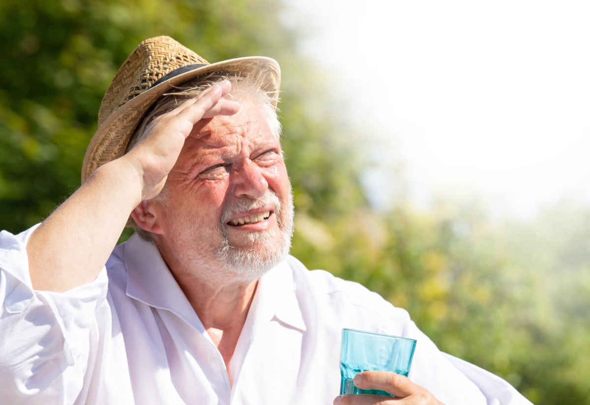 An elderly man sits outside in the great heat. He sweats and drinks mineral water.