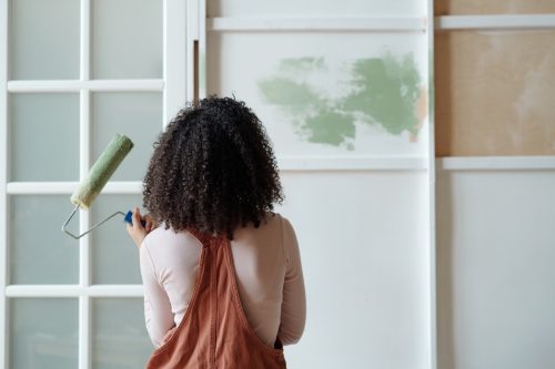 Rear view of young female owner of cafe with paintroller in hand standing in front of wall while painting it in green color