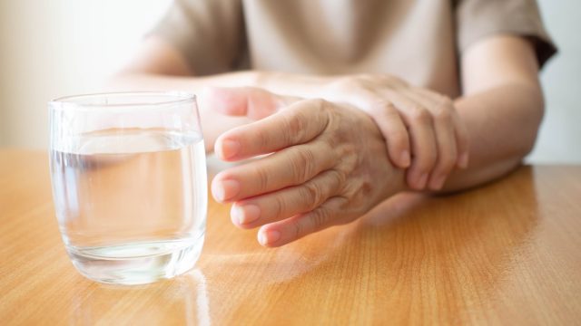 Elderly woman hands w/ tremor symptom reaching out for a glass of water on wood table. Cause of hands shaking include Parkinson's disease, stroke or brain injury. Mental health neurological disorder.