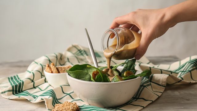 Pouring of tasty tahini from jar onto fresh vegetables in bowl