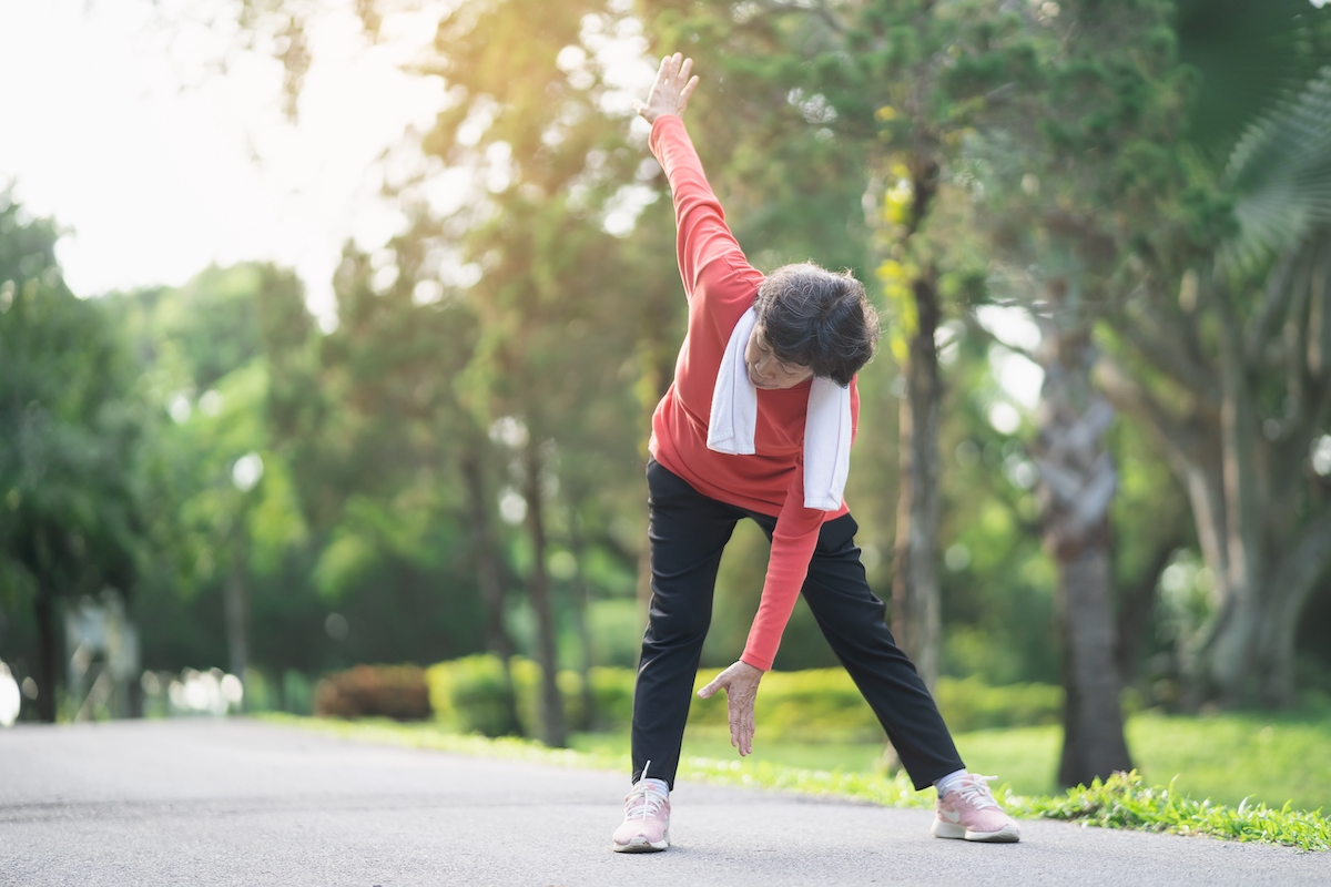 senior woman in black pants and a red top stretching to touch her toes in a park