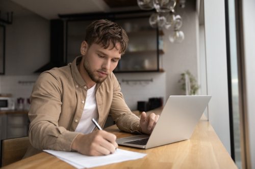 man handwriting list while working on laptop