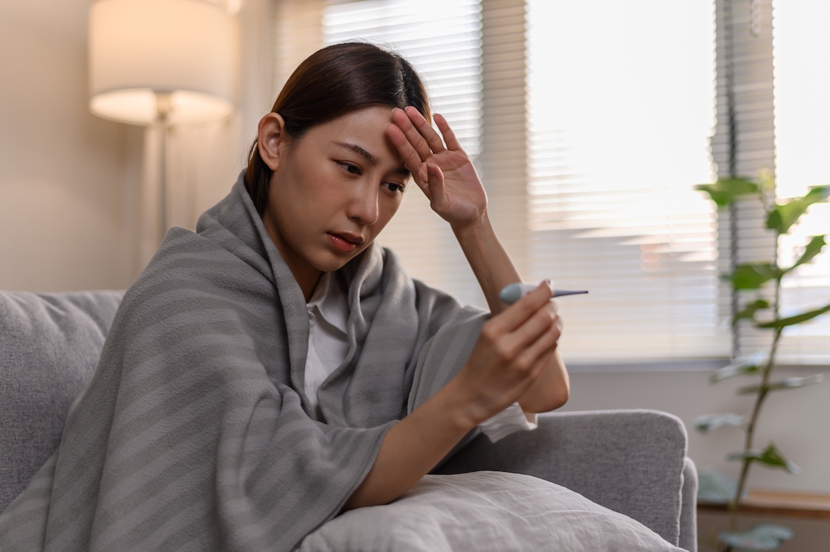 A sick young woman wrapped in a blanket on her couch, taking her temperature