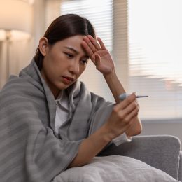 A sick young woman wrapped in a blanket on her couch, taking her temperature